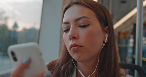 Close-up portrait of cute girl in headphones listening to music and browsing on smartphone in public transport. Woman looking out of a train window, thinking of something during her daily commute.