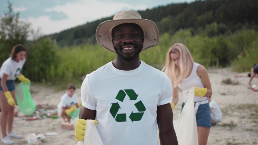 Modern ecology movement. Portrait of smiling afro-american handsome student picking up rubbish into plastic bag. Team of vigorous volunteers in eco T-shirts cleaning down the lake beach. Royalty-Free Stock Footage #1035821528