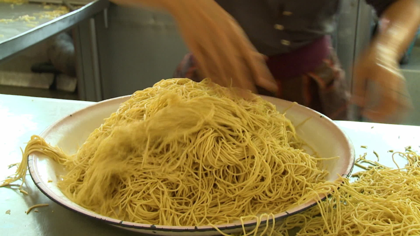 Handheld, medium close up shot of a person putting piles of noodles onto a large bowl.