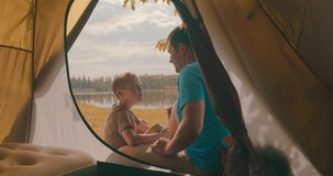 Happy traveler father and son sitting near tent camp around mountains lake under sun light enjoying the leisure and freedom. 4K slow motion video
