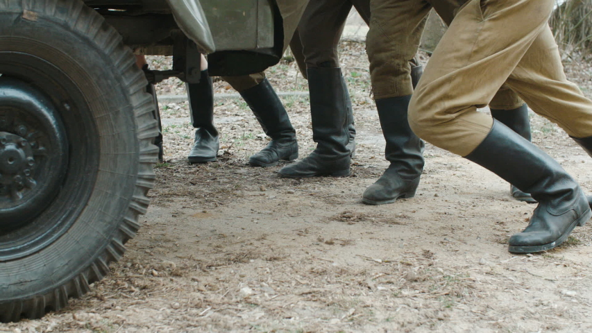 Close-up of boot soldiers in the form of world war II pushing a stalled vintage car. One man falls after being shot. The concept of evacuation in battle