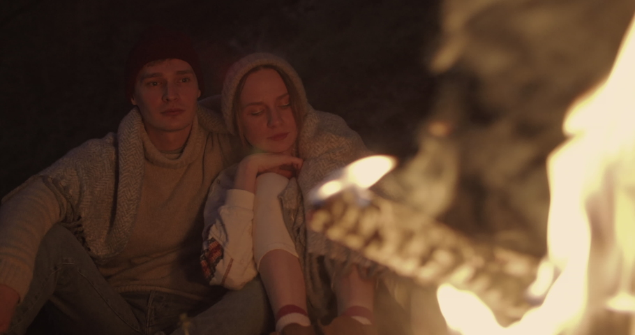 A young couple sits at night in the forest, embracing under a common plaid in front of a bonfire, talking and laughing.