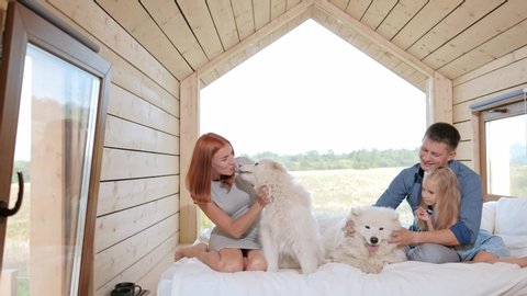 Young Caucasian family couple with baby daughter in a small modern rustic house with a large window. Lying on the bed, hugging, playing and looking out the window. Two Samoyeds. Weekend vacation