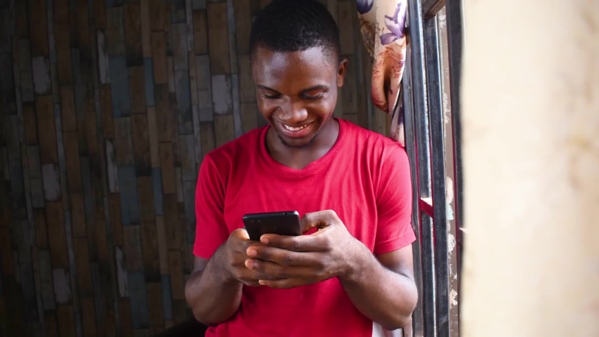 YOUNG BLACK AFRICAN MAN CHECKING INTERESTING CONTENT ONLINE ON HIS MOBILE PHONE. NIGERIAN GUY EXCITED  CHATTING ON SOCIAL MEDIA  Royalty-Free Stock Footage #1035871589