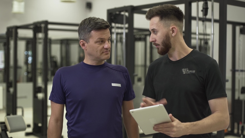Handsome professional fitness trainer with a tablet in hands explaining the plane of training for a sportsman. The man going to improve his body in the gym. Concept of sport, active lifestyle