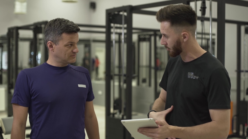 Young professional fitness trainer with a tablet in hands explaining plane of training for a new client. The man going to improve his press in the gym. Concept of sport, active lifestyle, healthcare