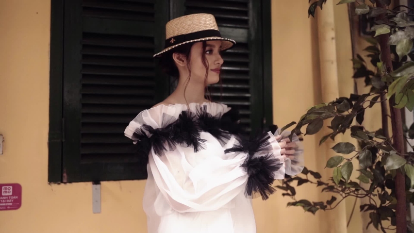 Beautiful and young fashion mestizo girl staying near the asian street with railway between old buildings and holding straw hat in hands. Journey concept in Vietnamese tourist destination in Hanoi. 