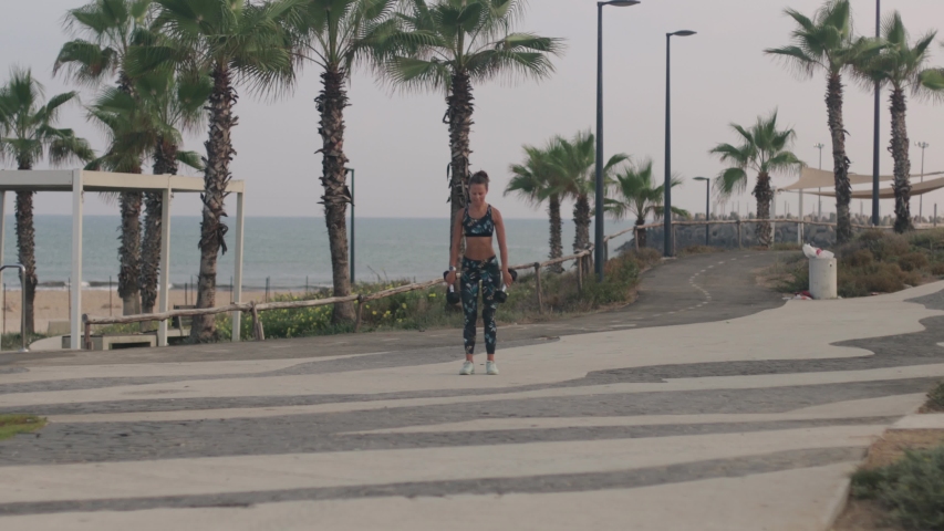 Young athletic woman lifting dumbbell weights, as part of her morning workout at the beach promenade.