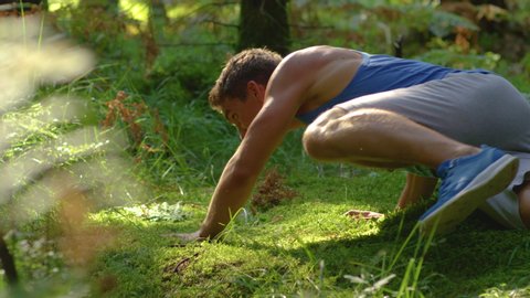 SLOW MOTION, CLOSE UP: Fit male jogger running through the woods trips and falls onto the soft mossy ground. Athletic young man stumbles while jogging through the peaceful sunlit forest in the summer.
