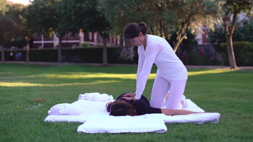 Shiatsu massage, shiatsu treatment. Wide shot of a masseuse pressing and massaging the back and the shoulders of a young beautiful woman at sunset in the park. Health care, wellness. Cine lens. Royalty-Free Stock Footage #1035898982