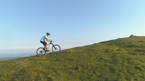 DRONE: Flying along an athletic man pedalling an e-bike up a steep grassy hill. Male tourist downhill biking in the Slovenian mountains riding his electric bicycle uphill on a sunny summer day.