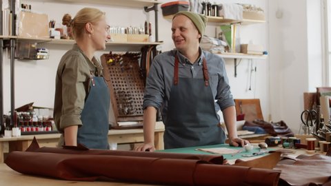 Medium shot of Caucasian man and woman standing leaning on worktop in leather workshop, talking then looking at camera and smiling