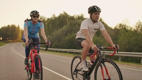 A man and a woman on bicycles ride down the road at sunset together in slow motion. The couple travels by Bicycle. Sports Cycling helmets