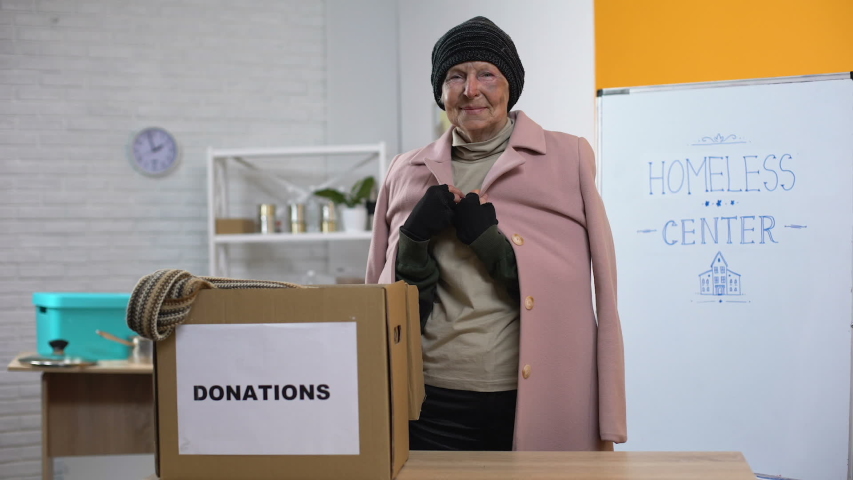Happy old homeless woman looking camera, donations box table, humanitarian aid