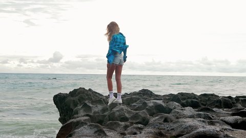 Young woman in modern outfit and checkered shirt walking on a large rock on the ocean. Tourist urban girl holiday. Summer vacation concept. Internet online freelance work break. Sport lifestyle.