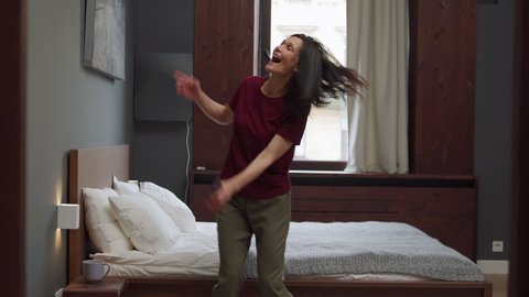 Tracking shot of happy mature woman dancing joyfully near bed in domestic bedroom or hotel room, celebrating good news with cell phone in her hand