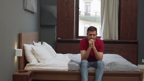 Depressed young man sitting on edge of bed at home or in hotel room and rubbing hands in anxiety. Nervous man looking at cell phone placed on bedside table awaiting for call suffering from addiction