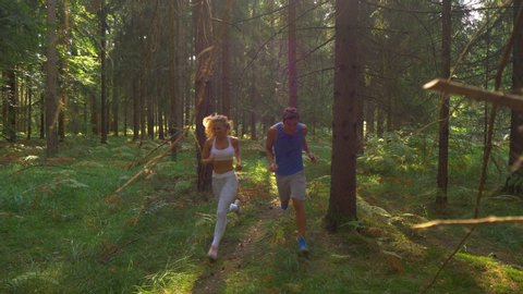 CLOSE UP, SLOW MOTION: Bright spring sunshine illuminates the scenic forest path for the two sporty trail runners. Cheerful Caucasian couple exercising and running around the idyllic green woods.