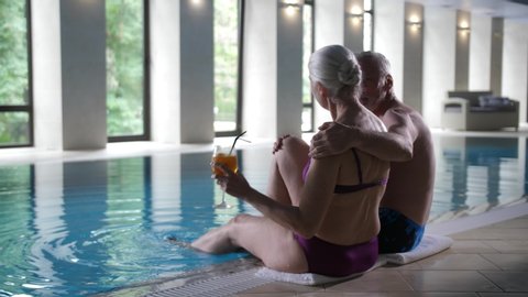Back view of loving mature husband and wife sitting on pool side, woman drinking cocktail, man hugging her gently. Happy aging couple enjoying vacation in luxury spa hotel, wealthy retirement concept
