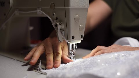 Tailor hands pulling white textile under presser foot of sewing machine. Working by light of built-in hardware lamp. Selective focus. Cutting off threads. Lace with sparkles on blurred foreground