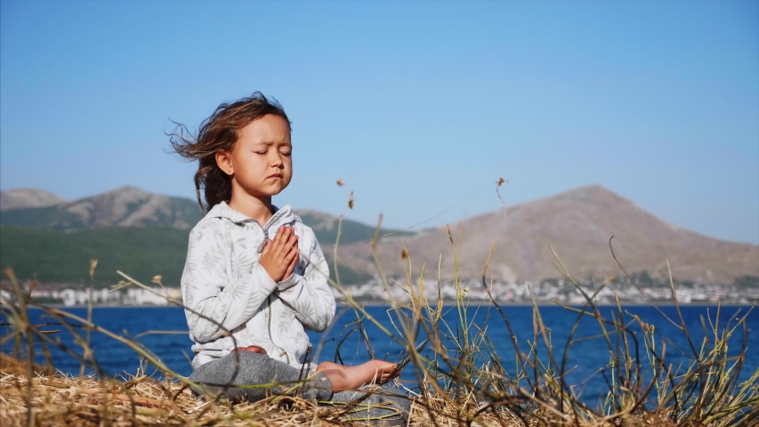 Cute little child gurl meditating alone in lotus pose at lake shore with namaskar gesture in windy weather