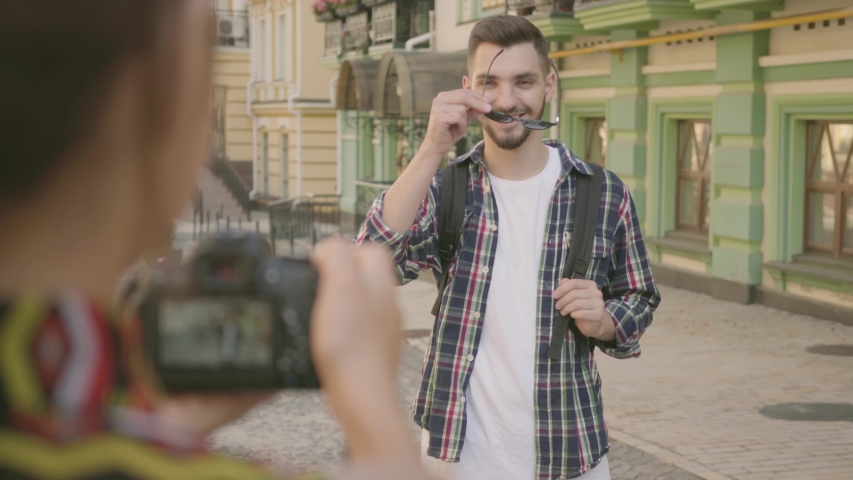 Female photographer taking a photo of the man in the street of an old city with beautiful architecture. Tourist takes picture of her friend. The funny guy posing in front of camera