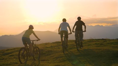 Three friends ride their electric bicycles up to the edge of a grassy hill and high five at sunset. Mountain bikers celebrate at sunset after a successful mountain biking journey in scenic Slovenia.