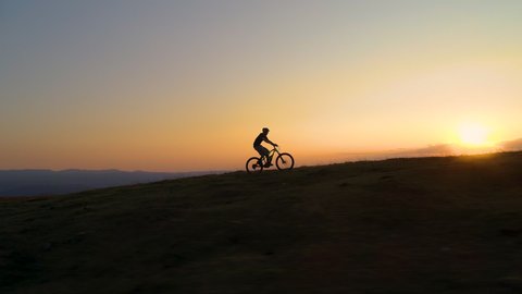 DRONE, SILHOUETTE, SUN FLARE: Unrecognizable cyclist rides an electric bike uphill at golden sunset. Breathtaking shot of an active tourist enjoying a scenic ride in the mountains on an e-bicycle.