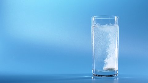 Effervescent Tablet Falling In Water. Pill Falls And Dissolves With Bubbles. Effervescent Tablet In Glass Of Water On Blue Background. Shot Of Effervescent Pill Dissolving In A Glass Of Water.