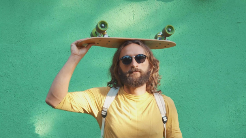 Portrait of a bearded long hair man dressed like hipster with black sunglasses in yellow t-shirt and organic backpack. He take a little wood skateboard on his head green wall background.  Royalty-Free Stock Footage #1035972440