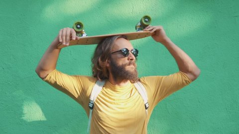 Portrait of a bearded long hair man dressed like hipster with black sunglasses in yellow t-shirt and organic backpack. He take a little wood skateboard on his head green wall background. 