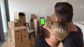 A moving couple looks at a smartphone with green screen in an empty apartment, a pile of cardboard boxes in the background