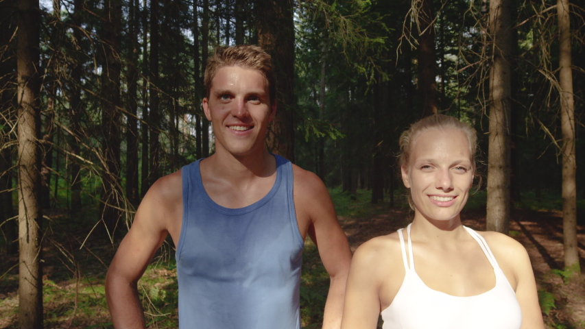SLOW MOTION, PORTRAIT, CLOSE UP: Smiling training partners stand in front of a forest after an intense jog in the wilderness. Smiling woman and her happy boyfriend look at the camera after a fun run.