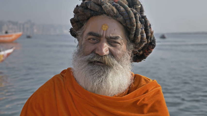 A bearded old or elderly male sadhu or Hindu priest wearing traditional clothes is sitting next to the Ganges River and looking at the camera while chanting a mantra or prayer in Varanasi