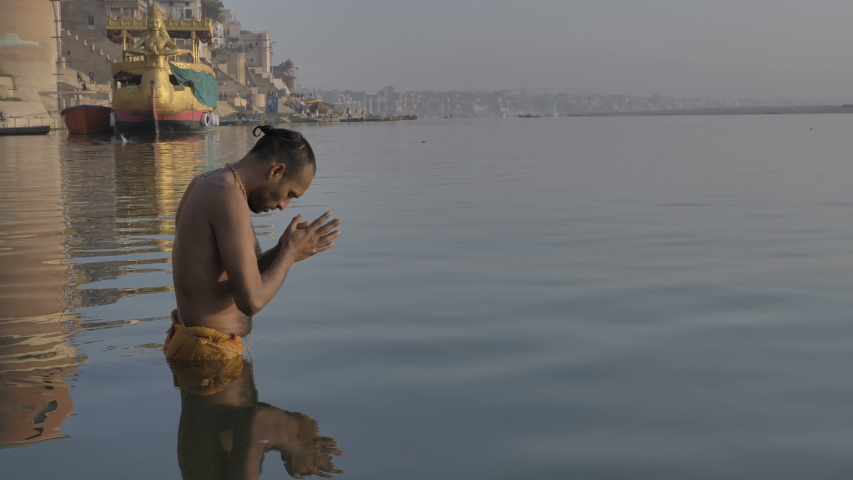A Hindu priest or pandit standing in the water on the banks of the Ganges, bowing before the Sun in the morning and offering Suryanamaskar, and praying to the morning sun on the banks of  Varanasi. Royalty-Free Stock Footage #1035982790