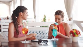 Two attractive girlfriends enjoying cocktails and watching photos on a smartphone in restaurant on the seashore in summer weekend. Friendship concept. Video shot of people on vacation in 4K definition