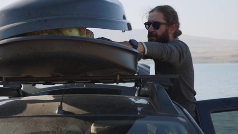 Young bearded man taking out a camping equipment from car roof box and throwing it on the ground, side view. Traveler is came to the lake and preparing for camping.