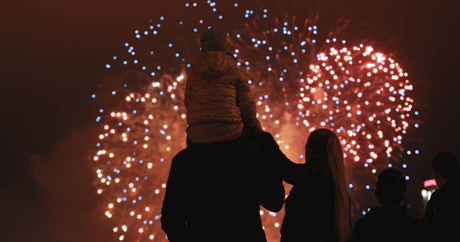Happy parents with a small child look at the sky and enjoy the party. Silhouette of people watching explosive and colorful fireworks in the evening sky. Holiday event and celebration. People have fun.