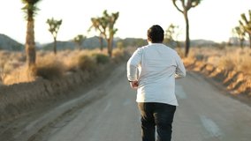 A millennial guy running alone in the rural dirt road towards the sun and running back towards the camera. wearing fashionable clothes. Slow motion video. 