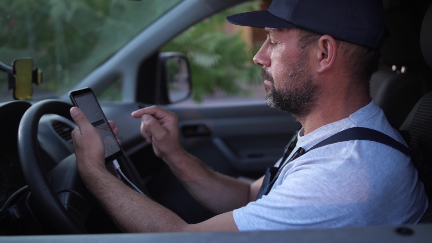 Worker of delivery service in uniform using cellphone navigator app setting route to client while sitting in car. Adult male in cap and overalls using smartphone to check route before delivering order Royalty-Free Stock Footage #1036013981