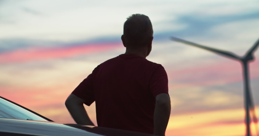 Back view of man enjoying summer pink sunset over large wind turbines spinning generating energy. Eco-friendly wind power station. Royalty-Free Stock Footage #1036016018