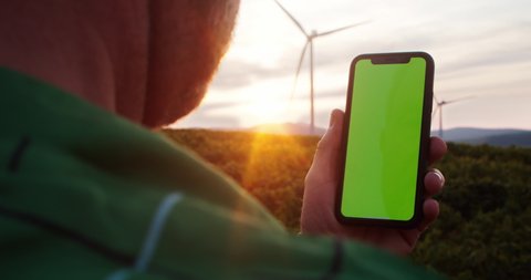Lviv, Ukraine - 15 August, 2019: Close-up of farmer using mock up greenscreen smartphone outdoor. Busy man with mobile phone in rural field with eco-friendly wind turbines spinning and sunset sky.
