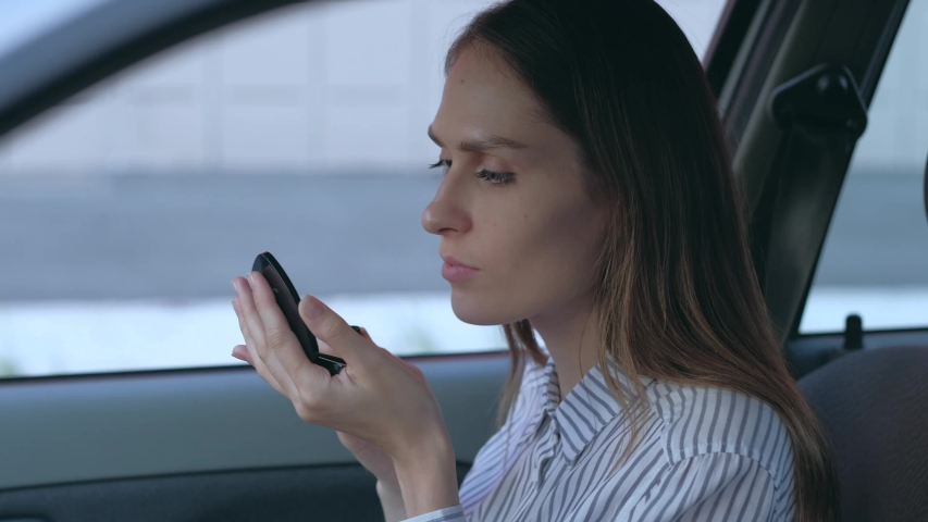 Female doing make-up, looking on herself at mirror and applying red lipstick on her lips into a car.