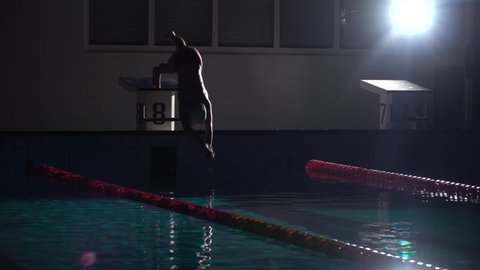 Female swimmer jumps off starting block and start swims in pool HD slow-motion video. Professional athlete training: dive and splashes water surface.