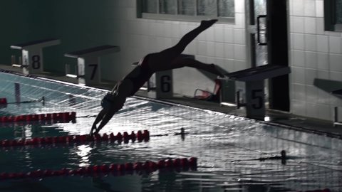 Female swimmer jumps off starting block and start swims in pool HD slow-motion video. Professional athlete training: dive and splashes water surface.