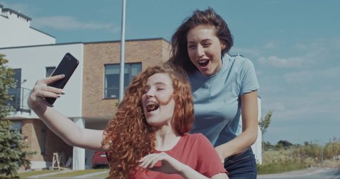 Two happy girlfriends spending time outdoors on lovely sunny day. Pretty girl riding a disabled invalid ginger cutie taking selfies having fun activity in the street.