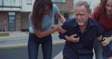 Portrait of stressed adult man feeling sharp chest pain as risk of heart attack in the street. In background two helpful girls running raising him up leading to the hospital.