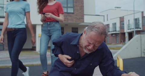 Mature man feeling sudden symptoms of heart-attack touching his chest falling down on ground. Indifferent young girls walking passing by without a help.