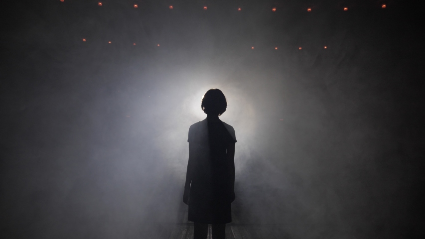 elegant silhouette of alone girl in short dress standing against bright spotlight in dark studio slow motion Royalty-Free Stock Footage #1036045151
