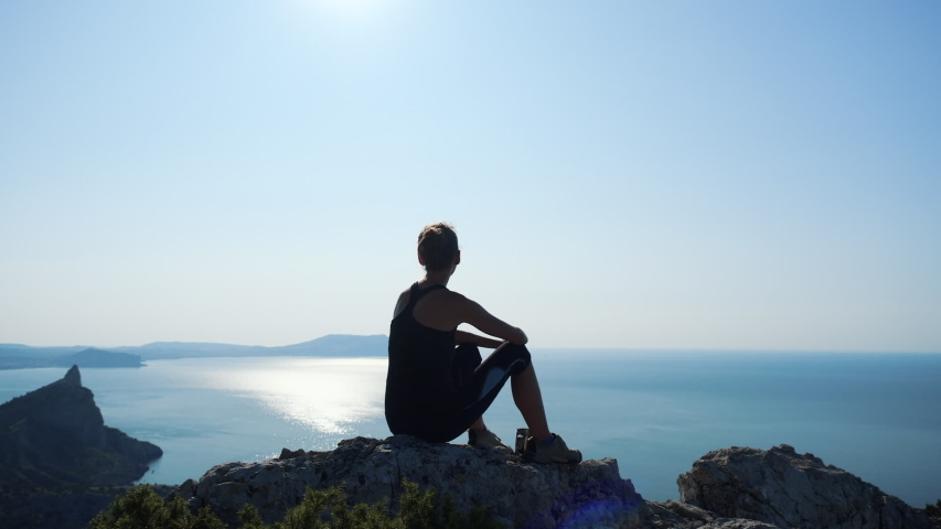 Young inspired woman sitting on the top of a mountain above the sea against beautiful blue sky. Silhouette of a happy hiker woman sitting on the summit and enjoying fantastic view from a hight.